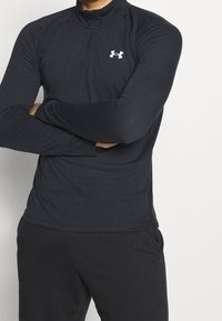 Black long-sleeve athletic top with a half-zip collar, featuring textured fabric and a white logo on the chest. Paired with black pants.
