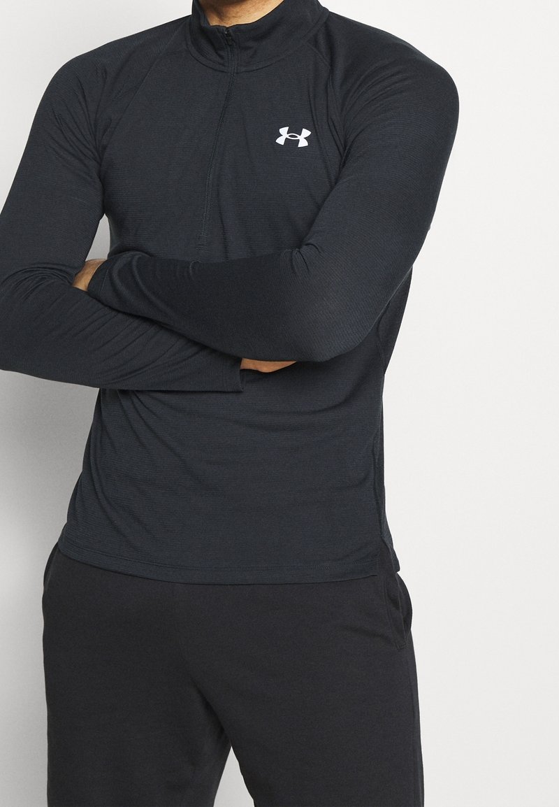 Black long-sleeve athletic top with a half-zip collar, featuring textured fabric and a white logo on the chest. Paired with black pants.