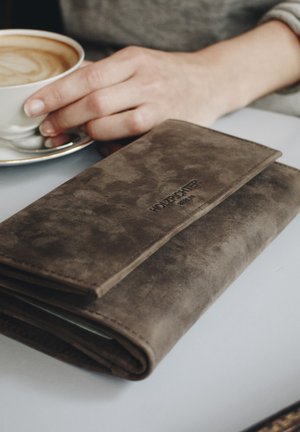 Brown suede wallet with a smooth texture, rectangular shape, featuring an embossed logo and a flap closure, positioned beside a coffee cup.