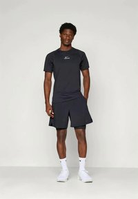 Mees seisab mustas Nike Pro sportlikus särgis ja lühikeses püksis, valgetes Nike sokkides ja valgetes spordijalatsites, lihtsa tausta ees.