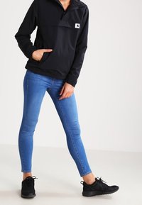 Sort zip-up jakke med en forlommer, sammen med blå skinny jeans og sorte sportsko. Glat stof, afslappet design, minimal branding.
