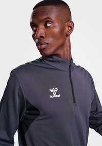 Grå idrotts zip-up topp med svarta sidopaneler, texturerad tyg och vit "hummel"-logotyp på bröstet. Slim fit och hög krage design.