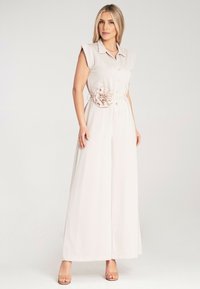 Jasno beżowy kombinezon z kołnierzykiem, zapinany na guziki z przodu, z krótkimi rękawami. Posiada akcent w postaci materiałowego kwiatu na pasku w talii oraz szerokie nogawki.