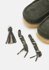 Green suede loafers with beige rubber soles, accompanied by green and beige shoelaces and a green-and-beige tassel keychain.