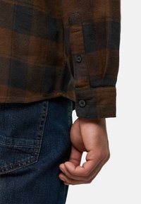 Brązowa i czarna koszula flanelowa w kratkę z zapiętymi mankietami, ukazująca teksturowany materiał obok niebieskich dżinsów i ręki w luźnej pozycji.