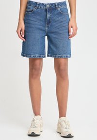 Medžiaginės bermudų šortai vidutinės mėlynos spalvos, su klasikiniu kirpimu, penkiomis kišenėmis ir šiek tiek suplėkusiu apvadu. Dėvimi su  šviesios spalvos sportbačiais.