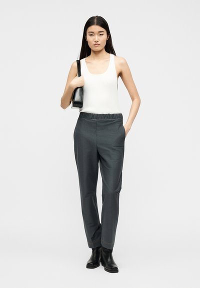 Γυναίκα που στέκεται, φοράει λευκό αμάνικο μπλουζάκι, σκούρο γκρι παντελόνι με ελαστική μέση, μαύρα μποτάκια αστραγάλου και κρατά μαύρη τσάντα ώμου.