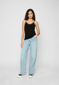 Svart tanktopp med blondekant, kombinert med lyseblå vide jeans. Modellen står med den ene hånden i lomma, iført svart og hvit joggesko.