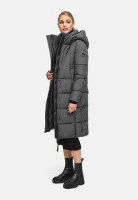 Lunga giacca imbottita grigia con cappuccio rimovibile, zip anteriore e tasche laterali. Indossata sopra un maglione nero e abbinata a stivaletti neri.
