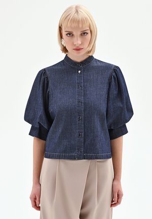 Camicia di jeans con maniche corte a sbuffo, chiusura frontale con bottoni e scollo con collo. Tessuto blu scuro con dettagli di cuciture sottili.