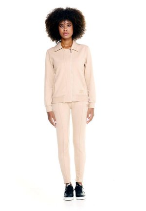 Une femme présente une veste beige zippée et un pantalon assorti, avec des baskets noires, sur un fond blanc.