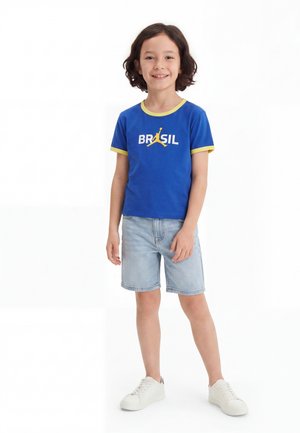 Παιδί που χαμογελά, φοράει μπλε μπλουζάκι «BRASIL» με κίτρινο περίγραμμα, ανοιχτόχρωμα τζιν σορτς και λευκά αθλητικά παπούτσια, στέκεται μπροστά σε λευκό φόντο.