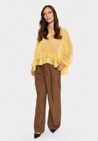 Yellow, tiered, sheer blouse with a V-neck, paired with brown, wide-leg trousers. The outfit features soft textures and a flowing design.