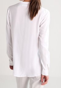 Blusa bianca a maniche lunghe con una texture liscia, collo rotondo e polsini con bottoni. Presenta un orlo leggermente curvo e una vestibilità su misura.