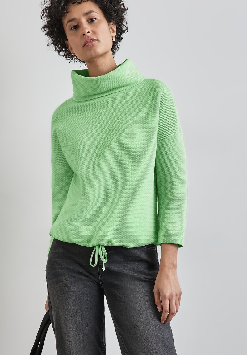 Grüner strukturierter Pullover mit hohem Kragen, Dreiviertelärmeln und einem Kordelzugsaum, kombiniert mit dunkelgrauen Jeans.