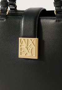 Sac en cuir noir texturé avec quincaillerie dorée. Présente une plaque logo carrée détaillant "Armani Exchange" en évidence sur le devant.
