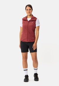 Gilet rosso senza maniche con zip frontale, abbinato a una camicia bianca, pantaloni corti neri e scarpe nere. Presenta un cappuccio e tasche con zip.