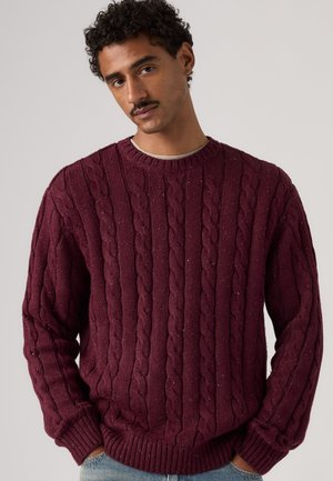 Homme aux cheveux bouclés et à la moustache portant un pull à torsades bordeaux et un jean bleu clair, debout devant un fond uni.