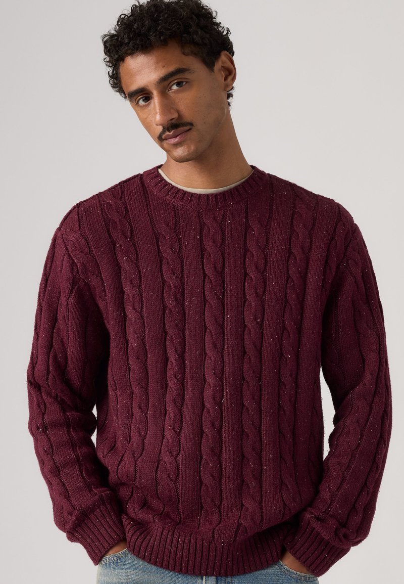 Homme aux cheveux bouclés et à la moustache portant un pull à torsades bordeaux et un jean bleu clair, debout devant un fond uni.