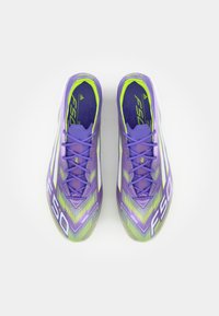 Purple soccer cleats with a textured surface, featuring a zigzag pattern in green and yellow accents. Laces and branding along the side.