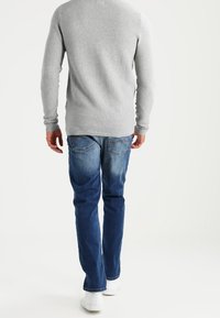 Homme marchant de dos portant un pull texturé gris clair, un jean bleu et des baskets blanches sur un fond blanc uni.