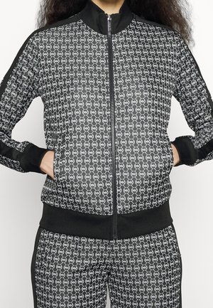 Kobieta w czarno-białej kurtce zapinanej na zamek oraz dopasowanych spodniach z powtarzającym się wzorem logo MK, ręce w kieszeniach kurtki.