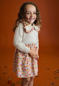 Lächelndes junges Mädchen mit lockigem Haar, das ein Blumenkleid mit einem gestrickten beigen Oberteil und einem Rüschensaum trägt, vor einem Hintergrund aus herbstlichen Blättern.