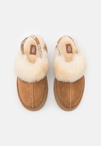 Chaussons en daim marron avec une épaisse doublure en fourrure crème et des brides arrière avec le logo UGG, vus de dessus sur un fond blanc.