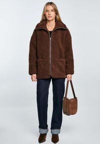 Braune Fleecejacke mit hohem Kragen, Reißverschluss vorne und zwei vorderen Taschen. Kombiniert mit dunklen Jeans und einer passenden braunen Tasche.