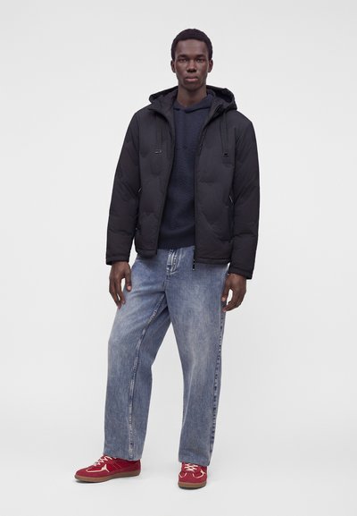 Jeune homme debout portant une veste à capuche noire, un pull sombre, un jean bleu clair ample et des baskets rouges sur un fond uni.