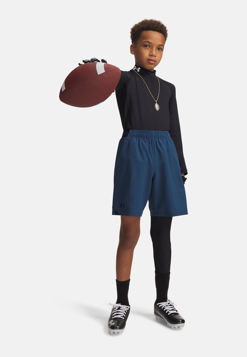 Garçon portant un t-shirt noir à manches longues, un short bleu et des crampons, tenant un ballon de football devant lui avec la main droite.