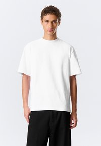 Homme aux cheveux courts et bouclés portant un T-shirt blanc uni oversize et un pantalon noir, debout devant un fond clair.