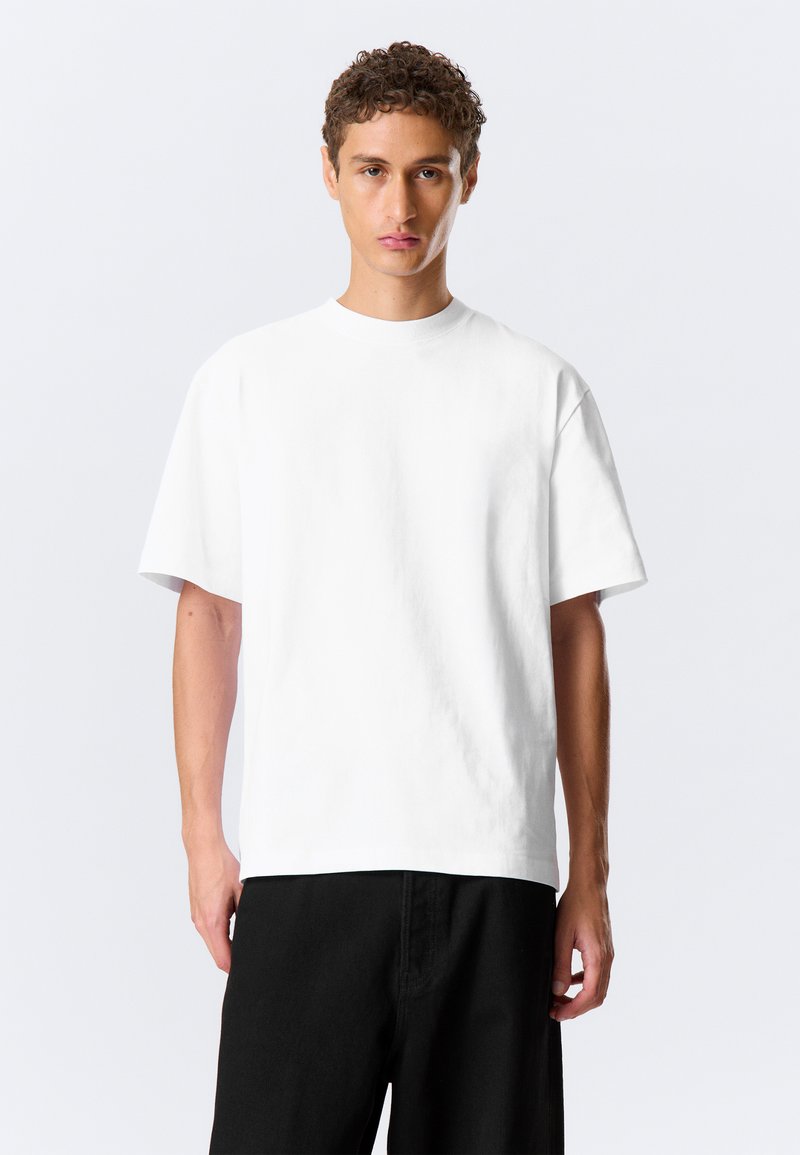 Homme aux cheveux courts et bouclés portant un T-shirt blanc uni oversize et un pantalon noir, debout devant un fond clair.