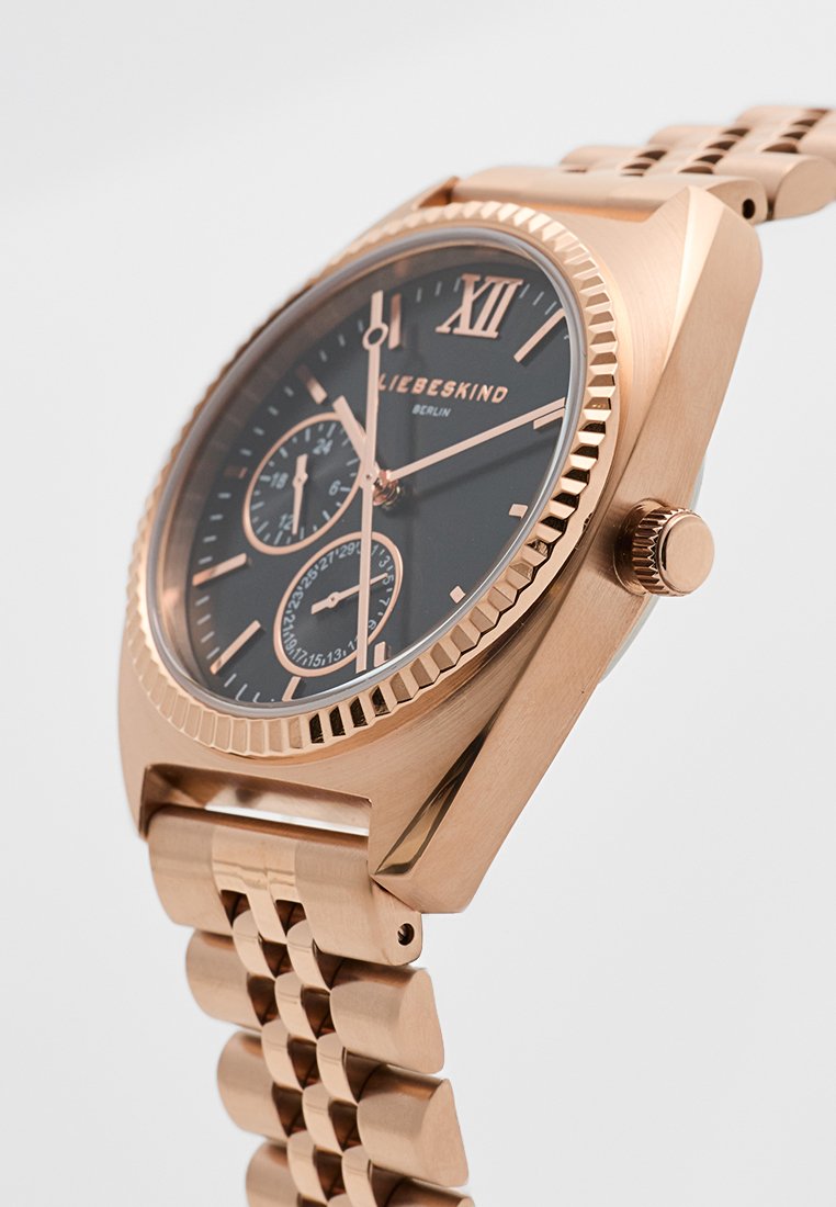 Reloj de metal rosa dorado con caja cuadrada, esfera negra, tres subesferas, número romano XII y banda de pulsera texturizada con diseño de eslabones.