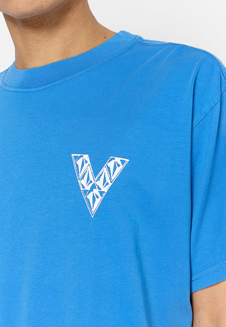 T-shirt en coton bleu présentant un design géométrique blanc en forme de "V" sur le côté gauche de la poitrine. Col rond et manches courtes avec une texture lisse.