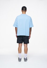 Svetle modré oversized bavlněné tričko v kombinaci s černými šortkami, nošeno s bílými ponožkami a černými teniskami. Jednoduchý design, bez vzorů nebo viditelných log.