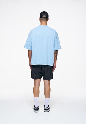 T-shirt oversized di cotone blu chiaro abbinata a pantaloni corti neri, indossata con calze bianche e sneakers nere. Design semplice, senza motivi o loghi visibili.
