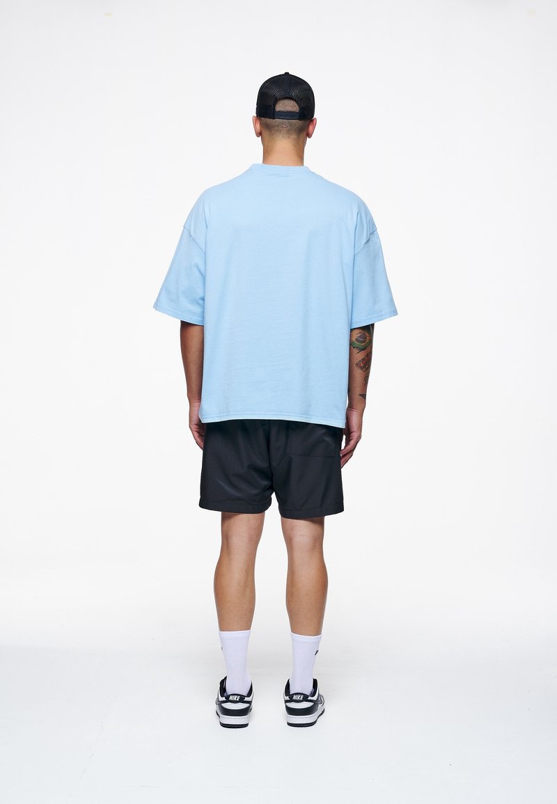Svetle modré oversized bavlněné tričko v kombinaci s černými šortkami, nošeno s bílými ponožkami a černými teniskami. Jednoduchý design, bez vzorů nebo viditelných log.