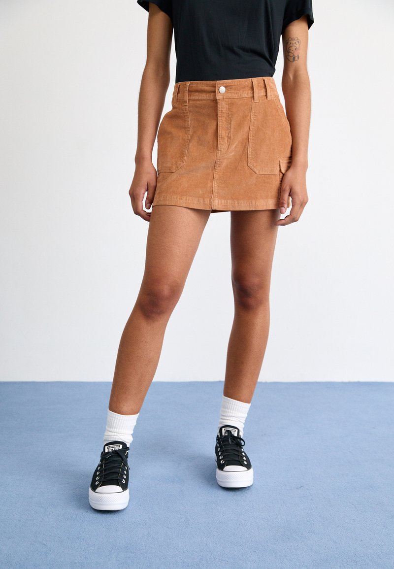 Bruine corduroy mini rok met voorzakken, hoge taille en knoopsluiting, gecombineerd met zwarte sneakers en witte sokken.