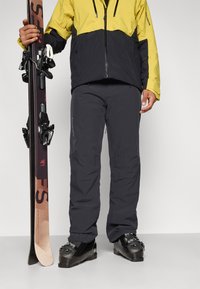 O fato de esqui apresenta uma jackete amarela e preta, calças cinzentas escuras e botas de esqui pretas. Inclui um esqui com as ligações encostado a uma perna.