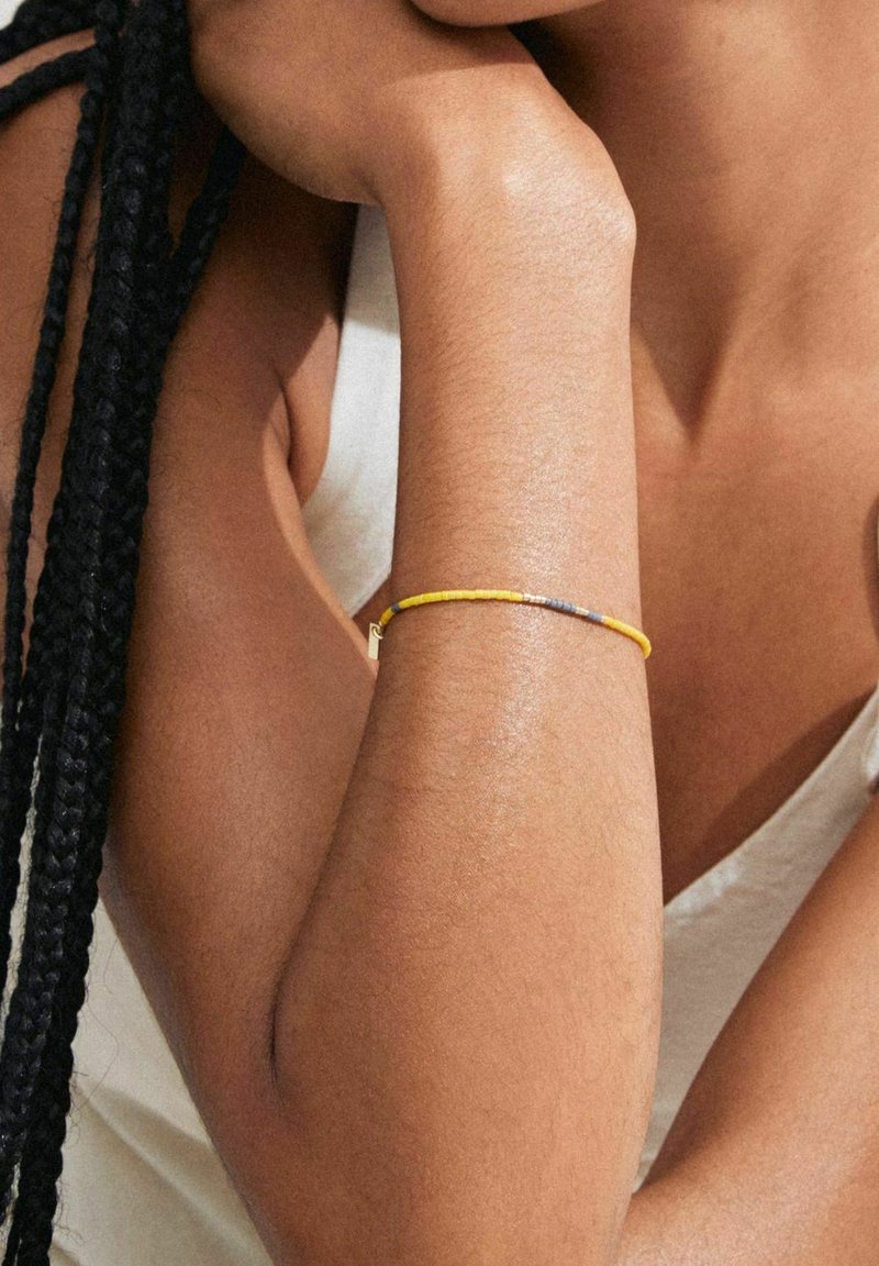 Bracciale sottile con perline gialle e grigie e un accento in hardware dorato, indossato su un braccio nudo con una leggera lucentezza.