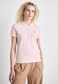 Lyserosa polo-skjorte med krage, fem-knapp placket og frontlogo. Stoffet er glatt og elastisk. Børt med beige bukser.