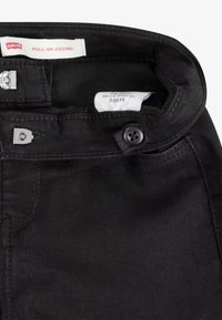 Jeggings negros de mezclilla elástica, con cierre de botón plateado y una cinturilla cosida con etiquetas. Textura suave.