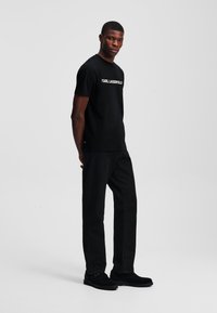 Juoda medvilnė crew-neck marškinėliai su balta užrašu "KARL LAGERFELD", derinami su juodomis kelnėmis ir juodais batais, prieš paprastą foną.