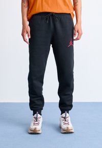 Person iført sorte joggers med rød logo, hvide og beige sneakers, og en orange skjorte, der står på en lyseblå gulv mod en hvid væg.