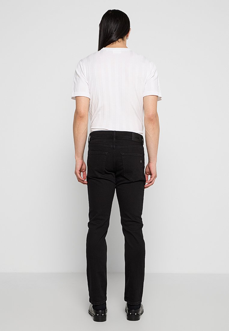 Jeans slim noirs avec poches arrière. T-shirt court à manches courtes en maille blanche. Le mannequin est de dos contre un fond uni.