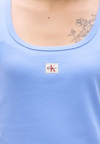 Ανοιχτό γαλάζιο ριμπ τοπ με στρογγυλεμένο λαιμόκοψη και ένα μικρό μπατζάκι με το λογότυπο Calvin Klein ραμμένο μπροστά. Λεπτή υφή, casual σχεδίαση.