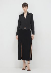Veste noire courte avec fermeture éclair à l'avant, associée à une longue jupe noire avec des fentes latérales et des talons aiguilles noirs à lanières. Texture de tissu lisse.
