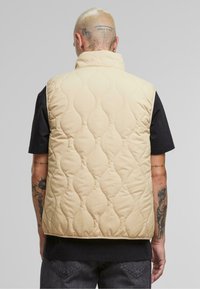 Padded vest with a beige quilted design, high collar, and armholes. Worn over a black t-shirt, showcasing detailed tattoos on arms.