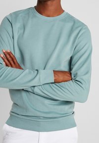 Sudadera de color verde azulado claro hecha de tela suave, con cuello redondo y puños acanalados. El diseño es sencillo con un ajuste holgado.